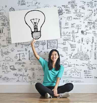 An image of a girl holding up a placard of a bulb. The background of the image has a number of graphs, equations, pie charts, etc. drawn on a white wall.
